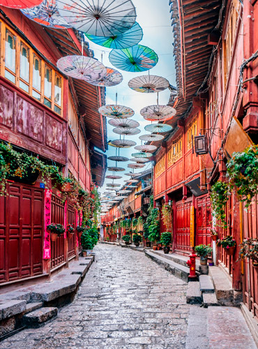 Un artesano mayor pintando a mano un fénix en un paraguas de seda junto a una joven tejiendo en un antiguo callejón adoquinado de una ciudad china tradicional, bajo un techo de sombrillas decorativas.